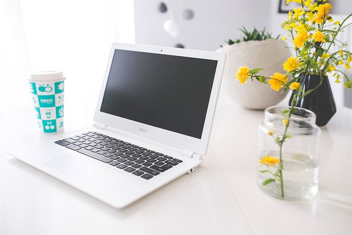 Clean white desk with a laptop, a takeaway cup and a small vase of yellow flowers