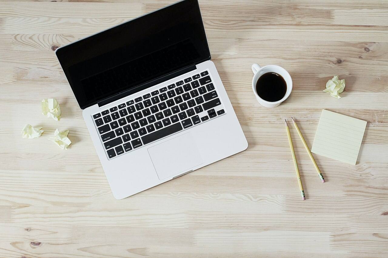 Top view of an open laptop, a cup of black coffee, two pencils and a sticky note on a light wooden desk