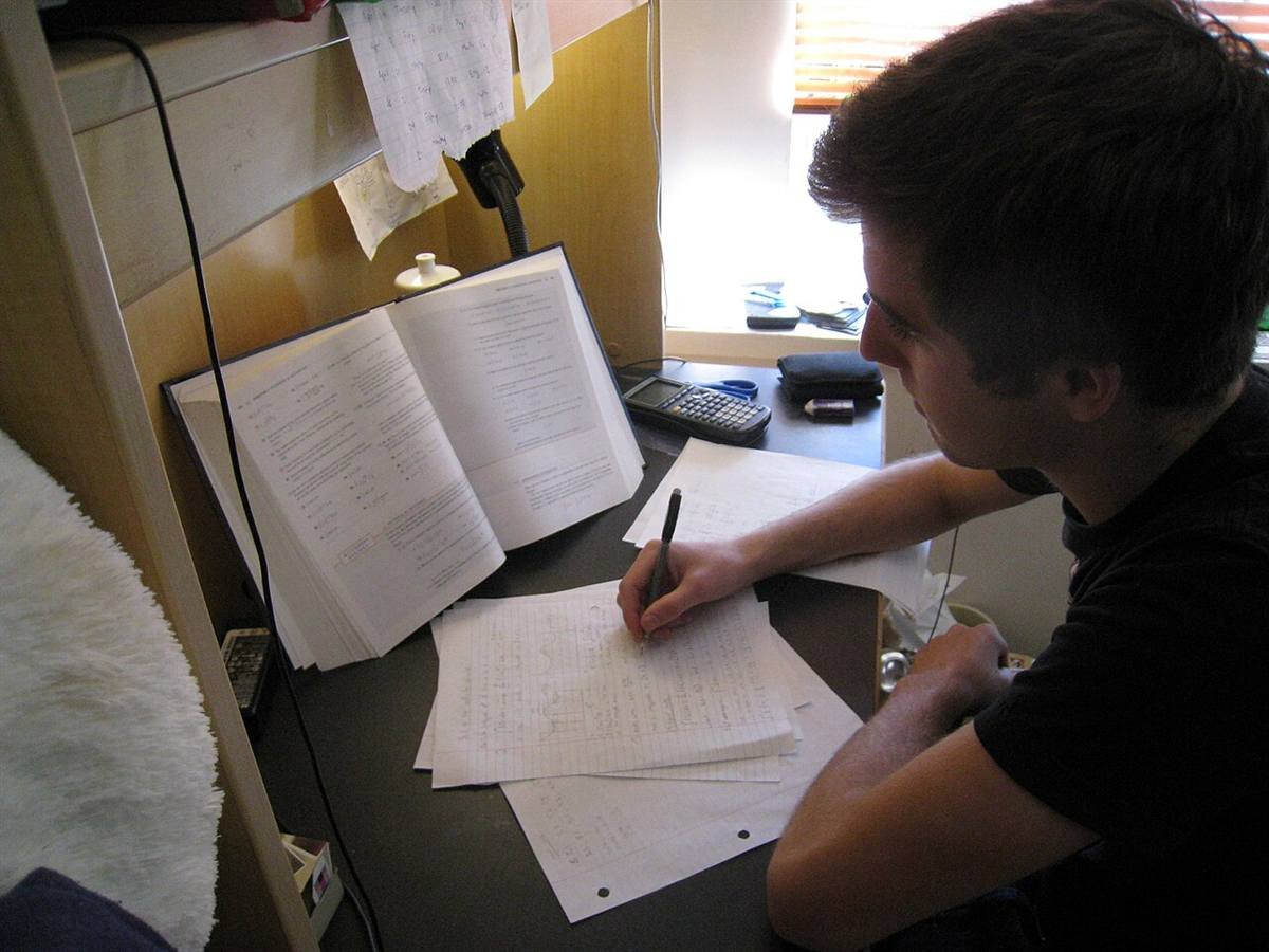 Student writing notes by hand at a desk, with an open textbook and a calculator beside him